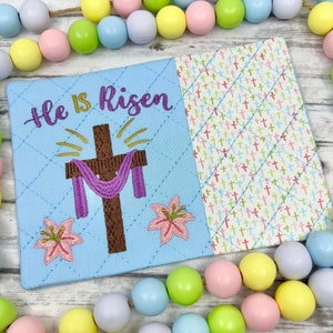 May include: A blue fabric Easter-themed pouch with a cross, lilies, and the text "He is Risen". The pouch is surrounded by a string of pastel-colored wooden beads.