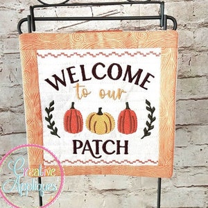 May include: A mini quilt hanging on a black stand with the text "Welcome to our Patch" and three embroidered pumpkins. The quilt is 8x8 inches and has a woodgrain pattern.