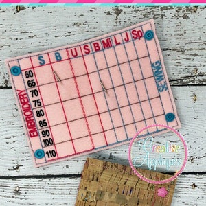 May include: A pink fabric needle minder with a grid for measuring embroidery sizes in inches. The grid is labeled with the letters S, B, U, S, B, M, I, L, J, Sp and the numbers 60, 65, 70, 75, 80, 85, 90, 100, 110. The words "EMBROIDERY" and "SEWING" are printed on the needle minder. The needle minder is sitting on a wooden surface.
