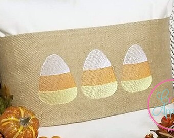 May include: A white pillow with a burlap accent featuring three embroidered candy corn designs. The pillow is sitting on a brown surface with two small pumpkins and a sprig of fall foliage.