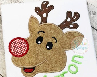 May include: A white t-shirt with an embroidered brown reindeer head with a red and white checkered nose. The reindeer has brown antlers with white polka dots. The name "Aaron" is embroidered in green below the reindeer.