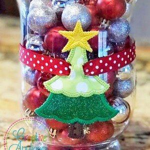 May include: A clear glass vase filled with red, silver, and white Christmas ornaments. A green felt Christmas tree applique with a yellow star is attached to the vase with a red and white polka dot ribbon.