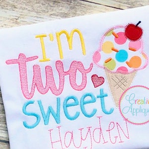 May include: A white onesie with the text "I'm two sweet Hayden" embroidered in pink, blue, and yellow. A pink heart and an ice cream cone with a cherry on top are also embroidered on the onesie.