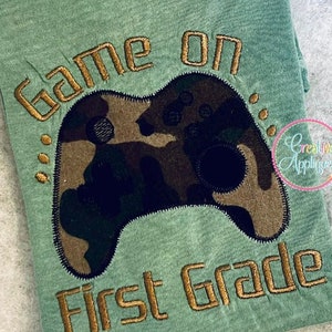 May include: Green t-shirt with gold embroidery that says "Game on First Grade" with a camouflage-patterned video game controller in the middle.