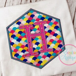 Könnte beinhalten: Ein weißes T-Shirt mit einem gestickten pinkfarbenen Buchstaben "H" in einer sechseckigen Form. Das Sechseck ist mit einem bunten Muster aus überlappenden Kreisen in den Farben Blau, Gelb, Pink und Weiß gefüllt. Der Text "Creative Appliques" ist in der rechten unteren Ecke des Bildes sichtbar.