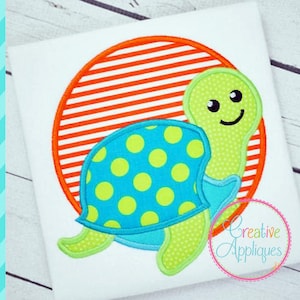 Może przedstawiać: Biała koszulka z nadrukiem z kreskówkowym żółwiem. Żółw ma zieloną głowę i nogi, niebieski pancerz z żółtymi kropkami, na tle pomarańczowo-białego koła w paski. Projekt zawiera tekst "Creative Appliques".