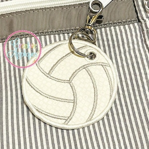 May include: A white volleyball-shaped tag with gray stitching, attached to a metal clasp. The tag is labeled "In The Hoop Tag" and is available in 3", 4", 5", and 6" sizes. The tag is set against a striped background.