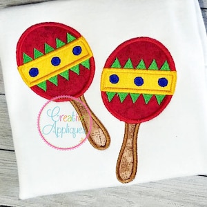 Puede incluir: Dos maracas rojas y amarillas con detalles verdes y azules. Las maracas están bordadas en una tela blanca.