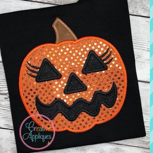 May include: Orange and black embroidered pumpkin with eyelashes and a toothy grin on a black fabric background. The pumpkin has a dotted pattern and a brown stem.