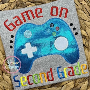 May include: Embroidered design on a gray shirt featuring a blue video game controller with the text "Game On Second Grade" in colorful letters. The design is available in four sizes.