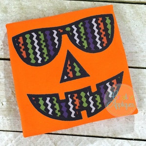 May include: Orange fabric with a black and white patterned jack-o'-lantern face wearing sunglasses. The sunglasses are also patterned with black, white, green, and purple.