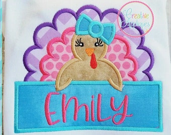 Puede incluir: Un pavo bordado en morado, rosa y marrón con un lazo azul. El pavo sostiene una pancarta azul con el nombre "Emily" bordado en rosa.