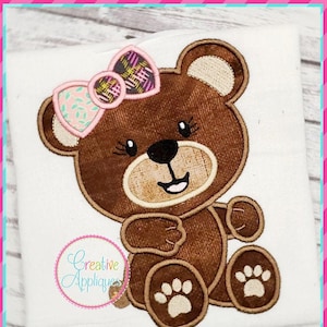 May include: A brown teddy bear with a pink and purple bow sitting on a white background. The bear has a happy expression and is wearing a bow with a floral pattern.