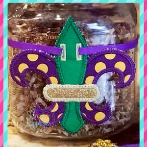 May include: Purple and gold Mardi Gras fleur-de-lis with yellow polka dots and a green center. The fleur-de-lis is attached to a clear glass jar with a purple ribbon.