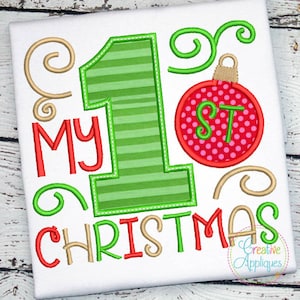 May include: A white onesie with the text "My 1st Christmas" in red and gold embroidery. The number 1 is green and striped. A red and white ornament with the letters "1st" is above the number 1.