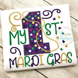 May include: White t-shirt with the words "My 1st Mardi Gras" in green, gold, and purple. The number 1 is purple with colorful polka dots and a gold beaded necklace. Confetti and swirls complete the design.