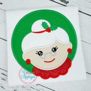 May include: Embroidered design of a smiling woman with white hair, wearing a red hat with a green holly sprig, red glasses, and a red collar. The design is surrounded by a green circle.