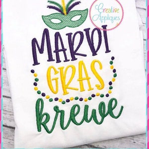 Op de afbeelding: Een wit T-shirt met een geborduurd ontwerp met een paarse tekst "Mardi Gras Krewe" met een groene en gele kralenrand en een groen en geel masker met paarse veren.