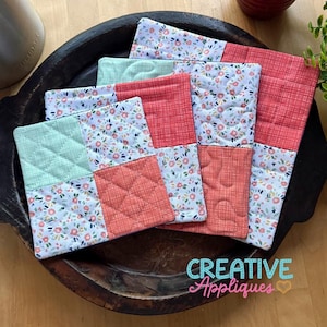 May include: A collection of quilted fabric coasters and placemats in a wooden bowl. The coasters and placemats feature a patchwork design with floral and geometric patterns in coral, mint green, and white. The text "CREATIVE Appliques" is visible.