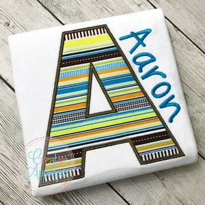 May include: A white t-shirt with a large, colourful striped letter A and the name "Aaron" embroidered in blue thread.