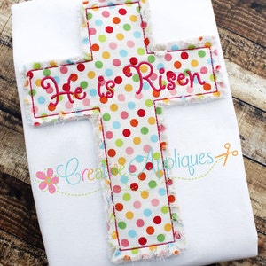 May include: A white t-shirt with a colorful polka dot cross applique. The cross has the words "He is Risen" embroidered in pink.