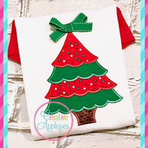May include: A white t-shirt with a red and green Christmas tree applique. The tree has a green bow on top and is decorated with red and white polka dots.