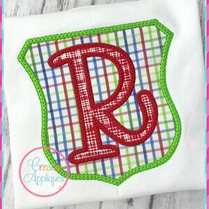 May include: A red letter R in a green and white plaid shield with a green border. The shield is on a white background.