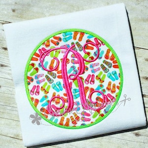 Może przedstawiać: Haftowany monogram litery "R" w kolorze różowym na białym tle z tkaniny. Litera jest otoczona kółkiem kolorowych klapek.