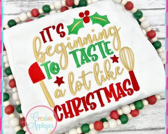 May include: White embroidery design with green holly, red berries, and gold text that reads "It's beginning to taste a lot like Christmas".
