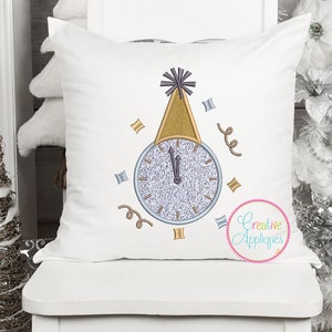 May include: White pillow with an embroidered design of a clock with a party hat on top. The clock face is silver with black hands and the hat is gold. The design is surrounded by confetti and streamers. The text "Creative Appliques" is embroidered in a circle in the bottom right corner.