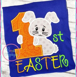 May include: A blue shirt with an embroidered design of a gray bunny holding a large orange number one. The text "1st Easter" is embroidered below the bunny.