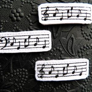 May include: Three white fabric patches with black embroidered musical notes and a treble clef. The patches are rectangular and have a stitched border.