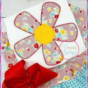 May include: A white t-shirt with a red and white floral applique in the shape of a flower. The flower has a yellow center and is surrounded by a red and white checkered pattern. The t-shirt is paired with a red bow and a blue and white patterned skirt.