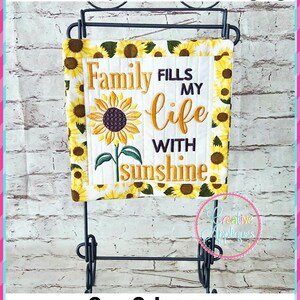 Op de afbeelding: Een mini-quilt die aan een standaard hangt met een zonnebloemdessin en de tekst "Family fills my life with sunshine". De quilt is 20,3 cm bij 20,3 cm.