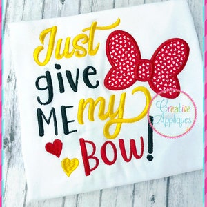 May include: White t-shirt with yellow, red, and black embroidery that reads "Just give me my bow!" with a red bow with white polka dots.