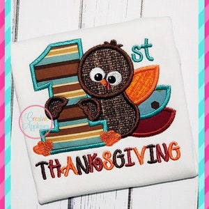 May include: A white onesie with a brown, orange, and blue turkey applique. The turkey is holding a large blue and brown number one. The text "1st" and "THANKSGIVING" is embroidered in brown, orange, and yellow thread.