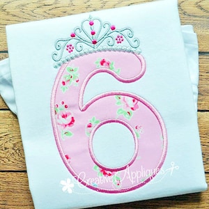 May include: A pink floral number six with a silver tiara on top. The number is embroidered on a white t-shirt.