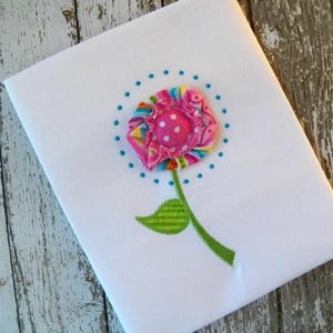 May include: A white fabric with a pink and blue flower embroidered on it. The flower has a pink centre with white polka dots and a green stem with a leaf.