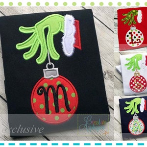 Könnte beinhalten: Drei bestickte Hemden mit einer grünen Grinch-Hand, die einen roten und grünen Weihnachtsbaumschmuck mit einem Buchstaben darauf hält. Die Hemden sind schwarz, weiß und dunkelblau.