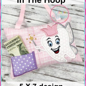 May include: A pink and white tooth pillow with a tooth fairy design. The tooth is wearing a pink crown and holding a magic wand. The pillow has a purple pocket with white polka dots. The pillow is 5 inches by 7 inches.