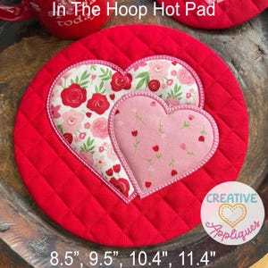 May include: Red quilted hot pad with two heart appliqués. The larger heart features a floral pattern with red and pink roses on a white background. The smaller heart has a pink floral pattern. The hot pad is available in 8.5", 9.5", 10.4", and 11.4" sizes.