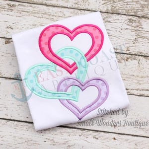 May include: Three layered hearts in pink, teal, and purple with white polka dots. The hearts are stitched together and have a white outline. The text "Stitched by Small Wonders Boutique" is at the bottom of the image.