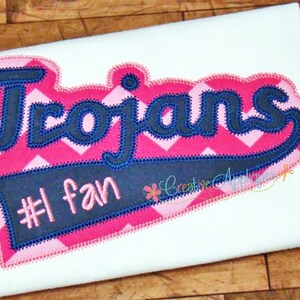 May include: Embroidered patch with the word "Trojans" in navy blue lettering on a pink and white chevron background. The patch also includes a navy blue banner with the text "#1 fan" in white lettering.