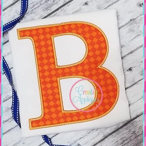 Könnte beinhalten: Orangefarbener und gelber Buchstabe B mit einem karierten Muster im Inneren. Der Buchstabe ist gelb umrandet und hat einen genähten Look.
