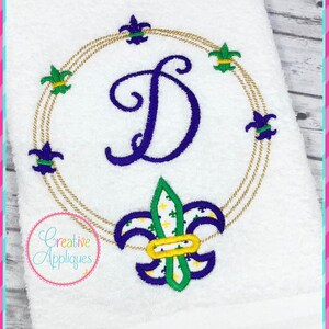 May include: White embroidered towel with a gold circle and fleur-de-lis designs. The letter "D" is embroidered in purple in the center of the circle. The text "Creative Appliques" is embroidered in the bottom left corner.