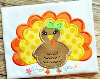 Puede incluir: Diseño bordado de un pavo marrón con plumas de cola de lunares amarillos y naranjas y un lazo verde. El pavo lleva un lazo verde.