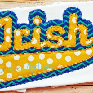 Könnte beinhalten: Eine gelbe Filzapplikation mit dem Wort "Irish" in einer fetten, abgerundeten Schriftart. Die Applikation ist mit einem blauen und grünen Chevron-Muster und weißen Punkten verziert.