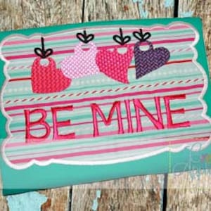 Könnte beinhalten: Stickerei mit fünf rosa, roten und violetten Herzen mit schwarzen Schnüren, die an einem türkisfarbenen Rahmen mit gestreiftem Hintergrund hängen. Der Text "BE MINE" ist in rosa auf dem türkisfarbenen Rahmen gestickt.
