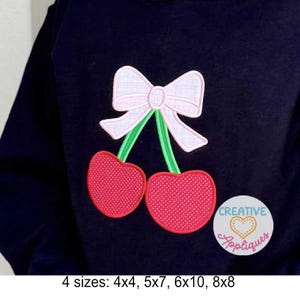 May include: A navy blue sweatshirt features a cherry design with a pink bow and two red cherries. The design includes green stems and is embroidered. The image also includes the text "Creative Appliques".