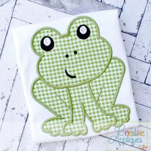 May include: White t-shirt featuring a green gingham frog applique. The frog has black eyes and a smiling mouth. The applique is outlined in green stitching. The shirt is a children's apparel item.